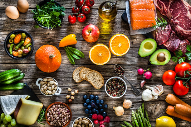 Overhead flat lay of diverse whole foods arranged on a rustic wooden table, including salmon, raw beef, avocado, oranges, pumpkin, tomatoes, berries, bread, nuts, cheese, vegetables, and olive oil.