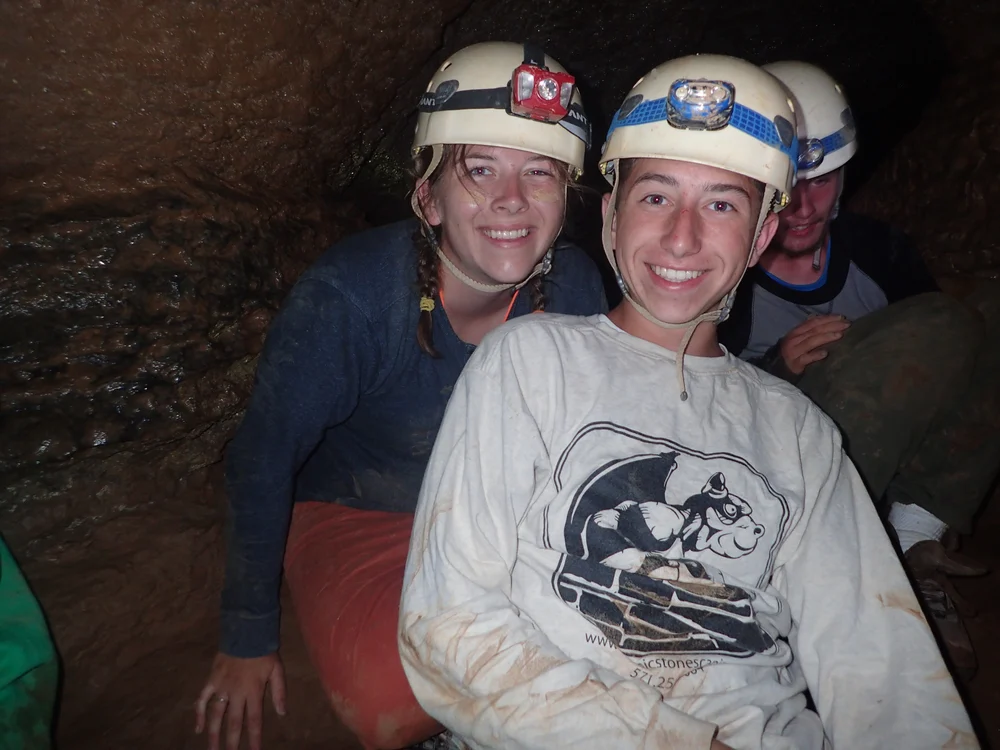 Smiling campers wearing helmets with headlamps pause for a photo deep inside a cave during a caving adventure, their clothes muddy from crawling through the underground passages.