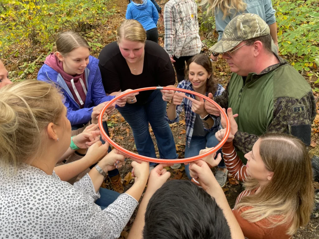A small group of adults collaborating on an outdoor team-building activity, working together to maneuver a hula hoop while standing in a circle in a wooded setting.