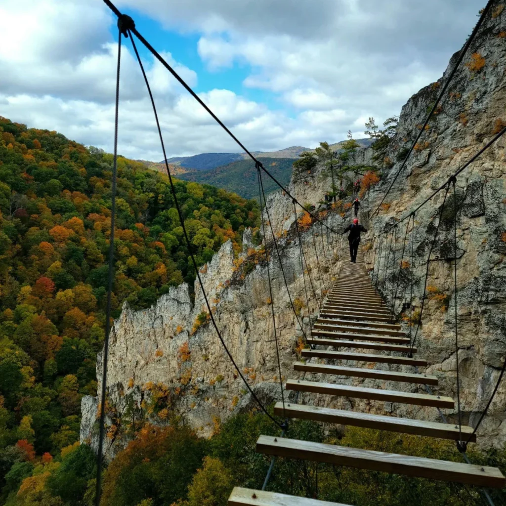 Adventurers wearing helmets cross a suspended wooden rope bridge along a dramatic rocky cliff face, with sweeping views of a mountain gorge filled with vibrant fall foliage below.